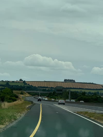 An eye-level, medium shot captures a view of a multilane highway curving through a rural landscape under an overcast sky in Hacklow, Ireland. The road, paved with dark asphalt, is divided by a bright yellow line on the left and a white line on the right, with a series of horizontal white lines marking a transition in the road on the far right. Several vehicles, including a black SUV and a silver sedan, are visible on the highway, with others in the distance. The road is bordered by grassy verges and shrubs. In the background, rolling hills are covered with a patchwork of green fields and a harvested golden field, with a cluster of trees on a distant ridge. A concrete overpass is visible spanning the highway in the mid-ground, and a tall, square sign structure stands to the left of the road.