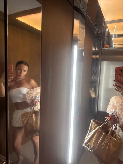 A woman takes a mirror selfie inside an elevator. She is wearing a white strapless top and a white ruffled skirt, accessorized with a pearl necklace and a gold tote bag. Her hair is pulled back in a bun. The elevator has mirrored walls and wooden accents. The image is taken from a low angle, and the lighting suggests it is daytime. The location is in the city of Zapadny, Russia.