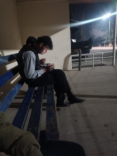 The image depicts an outdoor or semi-sheltered night scene, dominated by artificial lighting. In the foreground, a young male is seated on a worn blue wooden bench, intensely focused on a mobile phone held in both hands. His face is illuminated by the bright screen of the device, casting a soft glow. He has dark, curly hair and is wearing a light grey long-sleeved shirt under a dark (possibly black) vest, dark trousers, and dark dress shoes.
Behind this individual, another person is partially visible, also seated on the bench with their back towards the viewer. Only their dark-clad upper back and head are discernible. In the very bottom left corner, the lower body and legs of a third person are visible, suggesting they are also sitting nearby, possibly wearing a khaki or olive green jacket.
The bench itself is made of wooden slats, painted blue, and shows signs of wear, with patches where the paint has faded or chipped, revealing the natural wood underneath. Behind the bench, there is a light-colored wall or building facade that catches some of the ambient light.
The background opens up to an outdoor area. A large, dark vehicle, possibly a truck or trailer, is parked behind a metal railing or fence structure. Further back, a very bright street light on a pole emits a strong, star-like glare, casting long shadows and illuminating parts of the scene with a stark white light. Beyond the street light, sparse trees or bushes are visible in the distant darkness, against what appears to be unpaved or dirt ground. The ground immediately in front of the bench is a flat, dark surface, likely concrete or asphalt, with some faint markings or debris. The overall atmosphere is dim, with pockets of strong light from the phone screen and the distant street lamp creating high contrast. No text is visible in the image.