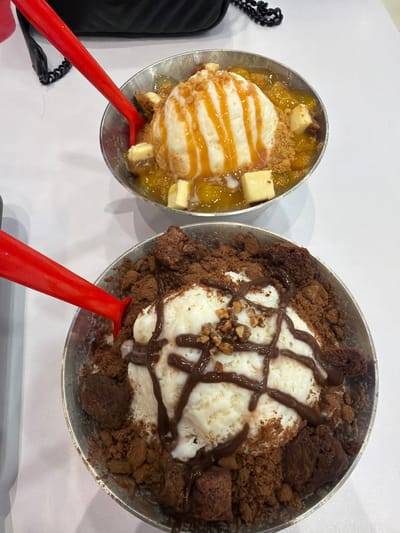Two metal bowls of shaved ice dessert are positioned on a white table. The bowl in the foreground contains a mound of white shaved ice topped with chocolate syrup and crushed nuts, surrounded by chocolate crumbles and brownie pieces. A red spoon is placed in the bowl, angled from the left. The bowl in the background holds a similar mound of white shaved ice drizzled with caramel sauce and topped with diced fruit and cookie crumbs, with a red spoon also inserted from the left. A black bag with a chain strap rests behind the bowls. The setting appears to be an indoor establishment, potentially a dessert shop or restaurant, with soft lighting. There are no individuals visible in the image. This scene could be located in Dagupan City, Philippines, given the context of the request.
