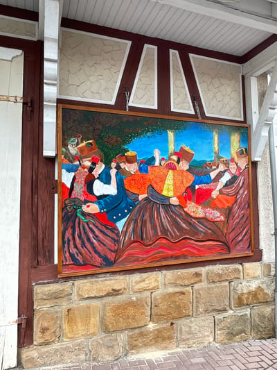 The image shows a large, framed mural depicting a scene of people in traditional attire dancing. The mural is mounted on a stone wall beneath a covered walkway with a wooden ceiling. The artwork features men and women dressed in brightly colored clothing, including hats and full skirts, with an overall folk art style. The background of the mural includes trees and a blue sky. The stone wall is constructed of irregularly shaped tan and brown stones. The walkway area is bordered by a white railing and has wooden beams supporting the roof. The area also features some architectural elements with a textured, cream-colored infill framed by dark red trim. A portion of a white wooden structure with a metal latch is visible on the left side of the image. The mural is located in the city of Willingshausen, Germany, known for its artist colony.