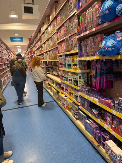 The image shows an aisle in a retail store, likely a toy section, located in Wrexham, United Kingdom. A blue floor and shelves stocked with various toys and games define the scene. Customers are browsing the merchandise, including adults and children. "Fashion Dolls" signage is visible. Shelves are filled with items like "K-POP idol" dolls, puzzles (Ravensburger), and games such as Monopoly Deal. Price tags are displayed on the shelves. A Powrmatic HVAC unit is mounted on the wall.