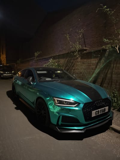 The image captures a vibrant teal-colored car, likely a performance vehicle such as an Audi, parked on a street or alleyway at night. The car is the primary subject, showcasing a distinctive iridescent metallic paint job that shifts from blue-green to green under the artificial lighting. A black racing stripe runs down the center of its hood. The front grille is a prominent black honeycomb design, and the car features sleek headlights and aggressive front splitter elements.
The front license plate is clearly visible and reads "C9 YAD".
Behind the teal car, another darker vehicle, possibly black, is partially visible, with its front end and a portion of its license plate ("EY60 XDX") discernible.
The setting appears to be an outdoor urban or suburban environment, characterized by a dark brick wall to the left, which has some climbing plants or overgrown vegetation on it. The ground surface is asphalt or tarmac. The overall darkness indicates it is nighttime, with artificial light sources illuminating the car and casting strong shadows on the ground and wall. The image is rotated 90 degrees clockwise from its natural orientation, presenting the car vertically. No people are visible, and no specific activity beyond parking is taking place. The location context provided is South Kesteven, United Kingdom, which is consistent with the visible UK license plate formats.