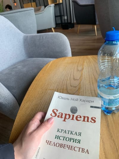 A person is sitting at a wooden table in a cafe, holding a copy of "Sapiens: A Brief History of Humankind" by Yuval Noah Harari, in Polish. The book is open to the cover, which features the book's title in large red letters, the author's name, and the words "Big Ideas" in small black letters. A partially filled plastic bottle of water with a blue cap rests on the table, adjacent to the book. The table is surrounded by gray upholstered chairs. The cafe is located in the city of Pyrzowice, Poland (coordinates: 50.471, 19.072). The scene appears to be indoors and well-lit. The person is not visible, only their right hand holding the book is.