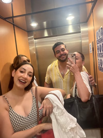 The image depicts four individuals inside an elevator, location in Chamilpa, Mexico as provided. The elevator interior features light brown wall panels with darker vertical accents, a reflective rear wall, and a side panel with white icons. Two recessed circular lights are visible on the ceiling.

In the foreground, a woman with dark hair and a broad smile wears a black and white patterned spaghetti strap top. She carries a reddish-orange shoulder bag and holds a white ruffled garment in her left hand. Behind her, partially obscured, is another woman with dark hair, wearing sunglasses pushed up onto her head.

To the right, a man with dark hair and a beard smiles, wearing a yellow and white striped short-sleeved shirt with a silver pendant necklace. His left arm is around the shoulders of a woman taking a mirror selfie. This woman, with light brown hair, holds a light-colored smartphone to her face, obscuring her mouth and nose. She has red nail polish on her left hand, which wears a ring on the ring finger. She carries a large black tote bag with white stitching in her right hand, while her left arm is positioned behind the man and the bag.