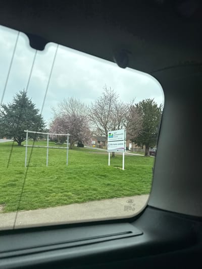 An exterior scene captured through a vehicle's rear window depicts a grassy area in Thrapston, United Kingdom, specifically near The Nene Centre. The sky is overcast.
In the immediate foreground, the dark grey interior trim of the vehicle's rear window is visible, with faint horizontal defroster lines and a small dark camera or sensor mounted centrally at the top edge. Below the window, a paved grey surface transitions to a meticulously maintained green lawn.
On the left side of the lawn, a light-colored metal frame structure stands, composed of multiple vertical posts and two horizontal crossbars, suggesting a support for signage or a sports goal. To the right, a white rectangular sign, mounted on two poles, is prominently displayed. The upper portion of the sign features the "North Northamptonshire Council" logo and text, including a phone number. The lower portion displays "the nene centre" in a stylized font, along with its website address (www.nene-centre.org.uk) and smaller logos such as "Active Nene" and "Healthy Thrapston".
The background features various trees; a large evergreen coniferous tree occupies the far left. Several deciduous trees, many bearing pink blossoms indicative of spring, are scattered across the midground. Behind these trees, residential-style buildings with light-colored facades and dark roofs are partially visible. On the far right, a dark-colored vehicle is partially obscured by trees. No individuals are present in the scene.