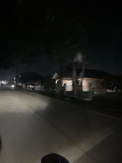 The image depicts a street scene at night in what is identified as Landuh, Indonesia. A paved road occupies the foreground, illuminated by the headlights of a vehicle, possibly a motorcycle given the low vantage point and the presence of what appears to be part of a handlebars or similar object in the very bottom of the frame. The road curves to the left, with a faint white line marking the edge.
On the right side of the road, there are several buildings. The closest building is a wooden structure with a light brown exterior, featuring visible horizontal planks. It has windows with what appear to be blinds or shutters. Further down the street, other buildings are visible, some with lit windows, suggesting habitation. A white fence runs along the front of some of these properties.
On the left side of the road, there are more buildings and trees. A single motorcycle, its headlight illuminated, is seen in the distance, traveling in the same direction as the viewer. Telephone poles and wires are also present, indicating infrastructure. The overall environment is dark, with the primary light sources being vehicle headlights and lights from the buildings. The image has a slightly blurred quality, possibly due to motion.