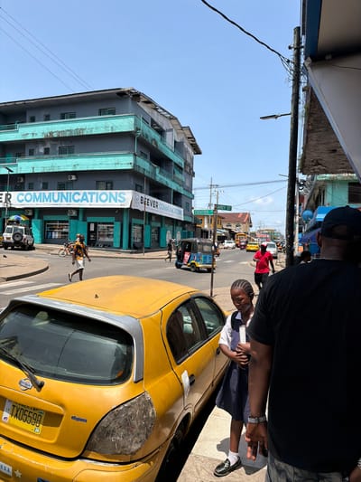 This image depicts a bustling street scene in Monrovia, Liberia, under a clear, bright blue sky during daytime. The perspective is slightly elevated, looking down a main thoroughfare.

In the foreground, a person in a dark cap and shirt is partially visible, with an arm extended. Next to them stands a young individual, likely a girl, wearing a school uniform (white shirt, dark skirt) and sporting braided hair. A prominent yellow Nissan car, possibly a taxi, occupies the right side of the foreground. It features a damaged front headlight area, and its license plate "TX05598" is clearly visible, along with the word "elegance."

The street is active with various forms of transportation and pedestrians. A blue tuk-tuk, a yellow bus, and other vehicles are seen. People walk along the street and sidewalks, including a man in a red shirt and another riding a bicycle. Utility poles are laden with a complex network of overhead wires.

On the left, a multi-story building with distinctive teal-colored balconies and window frames stands prominently. Spanning across the street or attached to a building is a large banner displaying "BEEVER COMMUNICATIONS" and "ER COMMUNICATIONS," alongside "Panasonic" and "RIGID." A green street sign in the mid-ground clearly marks "CENTER" and "CAREY ST." The overall scene captures the vibrant, everyday life of an urban setting.
