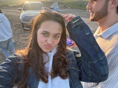 A selfie-style photograph shows two people outdoors. A woman with long brown hair, wearing a gray hooded sweatshirt and a blue denim jacket, is in the foreground. She is holding a pair of sunglasses in her hand. To the left of the frame is a man with a beard, wearing a striped shirt. A silver car is visible behind the woman. In the background, there is a grassy, hilly landscape, possibly in Hrazdan, Armenia, given the setting sun, terrain, and lighting. The lighting suggests the photo was taken near sunset.
