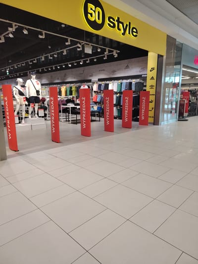 The image shows the entrance to a "50 Style" store in Wołomin, Poland, identified by the store's prominent yellow sign with black lettering. The store appears to be a clothing or shoe retailer, as evidenced by mannequins displaying outfits and racks of clothing visible inside. The entrance is partially blocked by several tall, red vertical banners with white text that reads "WYPRZEDAŻ," meaning "SALE" in Polish. These banners are spaced out, creating an aisle leading into the store. Inside the store, clothing is displayed on racks, and there are lighting fixtures on the ceiling. The floor in front of the store is tiled with large, light-colored square tiles.