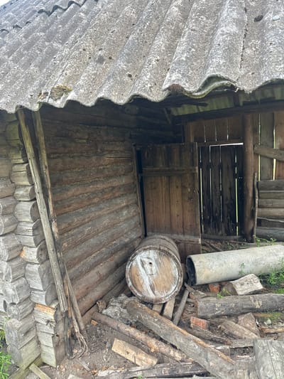 This image depicts the exterior of an old, dilapidated wooden structure, possibly a shed or an outbuilding, located in the Далёкаўскі сельскі Савет, Belarus. The building is constructed from rough-hewn logs, with a weathered, grey corrugated roof that shows signs of age and damage, including moss growth. The entrance to the structure is open, revealing a dark interior with wooden plank walls and doors. In front of the building, various objects are scattered on the ground, including a wooden barrel, pieces of pipe, bricks, and scattered logs and debris, suggesting neglect and disuse. The surrounding environment appears to be overgrown with vegetation, indicating a rural or semi-rural setting. There are no people or animals visible in the image.