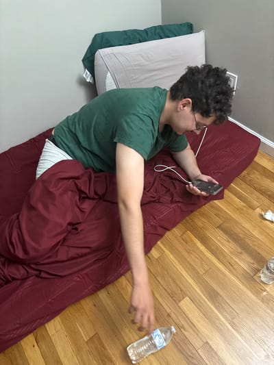 An overhead, diagonally oriented shot captures a young individual lying on a dark red sleeping bag or blanket placed directly on a light-colored hardwood floor in an indoor setting. The person, who has curly dark hair and wears glasses, is dressed in a green t-shirt and white striped underwear, and is partially covered by the red covering. They are positioned with their head resting on a light grey pillow, which is partially on a dark green pillow.
The individual is actively engaged with a smartphone held in their left hand, which is connected by a white cable, possibly a charger or headphones. The phone screen displays some unidentifiable white icons or text, possibly a dial pad or app interface. Their right arm is outstretched towards the floor where a clear plastic water bottle is visible. Another water bottle is situated near the top of the frame. The label on the closer water bottle partially reads "Great Value Purified Drinking Water". A crumpled piece of white paper is also on the wooden floor.
The background consists of a plain light grey wall. The person appears relaxed and focused on their device, with a slight smile on their face. The scene suggests a casual, laid-back atmosphere, possibly in a bedroom or dorm room in Franklin Township, United States of America. The time of day and weather are not discernible. No other significant text is clearly visible.