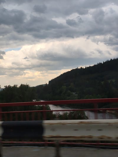 A view of a river valley from a bridge in Chusovskoye Urban Settlement, Russia. The river, reflecting the sky, is framed by dense green vegetation, primarily trees, on the left and a heavily forested hillside on the right. The sky is overcast with various cloud formations. The bridge structure includes a red painted rail and a weathered guardrail with alternating black and white sections. The photograph appears to have been taken from a moving vehicle.