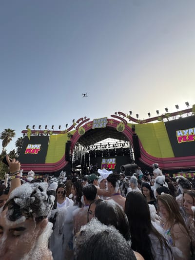 The image depicts a crowd of people covered in foam at a "Hype Fest" event in Grau I Platja, Spain. A stage with large screens displaying the "Hype Fest" logo is visible, and a drone flies above. The event is likely outdoors during the day. Palm trees are seen in the background. The focus is on the crowd experiencing the foam.