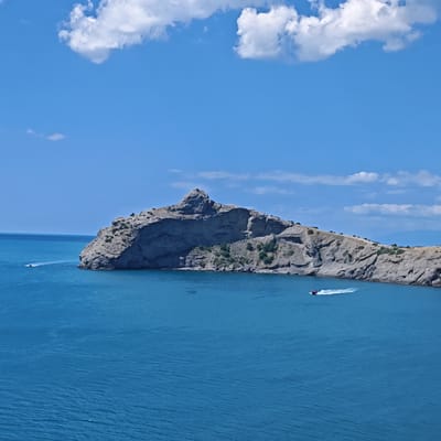 An elevated, wide-angle, full shot shows a body of bright blue water and a rocky, mountainous coastline under a clear sky with scattered white clouds. The coastline juts out into the water, with a prominent, jagged rock formation at its peak. Small patches of green vegetation are visible on the slopes of the cliffs. Two red and white speedboats are visible in the water; one is farther out to sea and appears to be moving away from the viewer, while the other is closer to shore and is creating a white wake as it moves to the right. The overall scene is serene and depicts a sunny day at the seaside, likely in a coastal area near Sudak, Ukraine, given the characteristic rocky formations and azure waters.