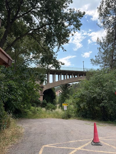 The image shows a view from below of the Aqueduct of Montanejos in Spain. The aqueduct is a concrete structure with rectangular openings between its support pillars. A streetlight is visible on the aqueduct's top. The sky is partially cloudy and seen through the aqueduct's openings and between the trees. A road runs along the right side of the image. A red traffic cone sits on the road next to yellow painted parking lines. A yellow sign in Spanish is near the side of the road that states "ENTRADA" and has some times listed. There are numerous trees and other vegetation throughout the image.