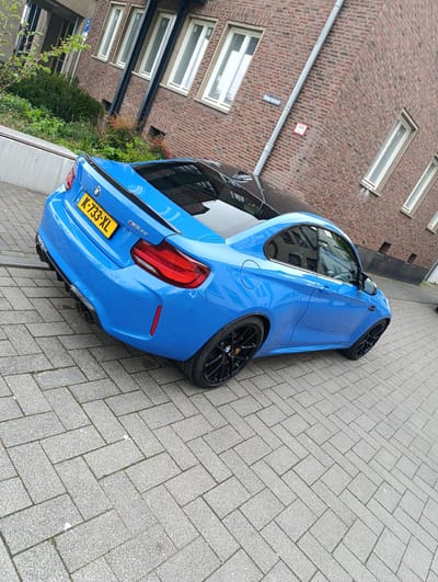 A blue BMW M2 CS is parked on a gray cobblestone street in Emmerich on the Rhine, Germany. The car's license plate is K-733-XL. The car has a black roof and black wheels. It is parked in front of a red brick building with white-framed windows. There is a street sign and building number on the brick building near the car. A concrete planter with green foliage is next to the building.