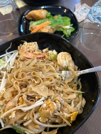 The image depicts a close-up view of a meal served on a tabletop within a restaurant setting in Louviers, France. In the foreground, a black bowl contains a serving of Pad Thai consisting of wide rice noodles, several cooked shrimp, bean sprouts, visible pieces of scrambled egg, and a topping of chopped peanuts. A silver metal fork is inserted into the noodles on the right side of the bowl, lifting some strands. In the background, slightly out of focus, a black rectangular plate holds two fried spring rolls (nems) accompanied by green lettuce leaves and shredded carrots. A small, peeled shrimp is also visible on this plate. To the left and right of the main food items, two clear glass tumblers are present on the textured, brownish-grey tabletop. A small white paper packet is partially visible behind the right glass. No individuals are depicted, but the presence of cutlery and prepared food indicates active dining.