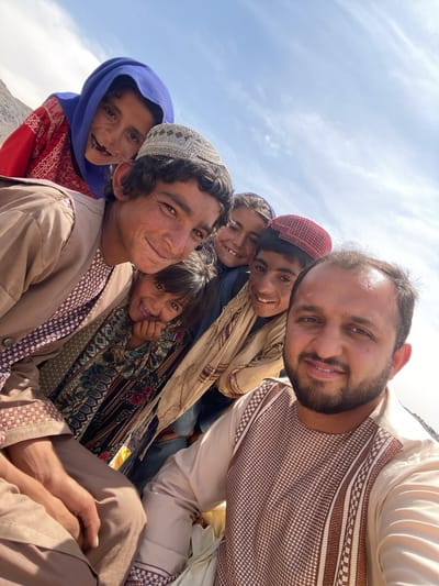 A selfie captures a group of Afghan individuals, including children and adults, in a likely mountainous setting near Ashoqah, Afghanistan. The foreground features a man with dark hair and a beard, wearing a light-colored shirt. Behind him are five children, all smiling and looking at the camera. One child wears a blue headscarf and a red dress, while another wears a cap. The clothing of the people suggests traditional Afghan dress. The backdrop appears to be a bright sky with visible mountain peaks.