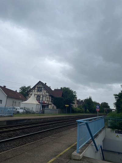 The image shows a train station platform in Hœrdt, France. The platform is made of asphalt and concrete, with yellow safety lines marking the edge. Two sets of train tracks run alongside the platform. To the left, there is a building with a white tent in front of it. The building is a traditional half-timbered house with a red tile roof. Above the tent, the building has signs that read "Tanz & Akiver" and "Pizzeria". There are cars parked in front of the building.

To the right of the platform, there is a set of stairs leading down to an underpass, with a blue metal railing. There are signs visible, including one that says "Passage Interdit" (No Passage) with a pedestrian symbol. The sky is overcast and grey, suggesting cloudy weather. Trees and other buildings are visible in the background.