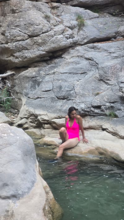 A full-body, portrait-oriented shot shows a woman sitting on a rock by a shallow body of water. She is wearing a neon pink one-piece swimsuit and has dark hair pulled back. One leg is extended into the water, with a dark shoe visible. The woman is looking down towards the water. The background consists of large, gray rock formations with some sparse vegetation. The water is clear with some small bubbles visible. The scene appears to be a natural outdoor setting, possibly a creek or pool within a rocky canyon.