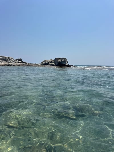The image captures a serene coastal scene with clear, turquoise water in the foreground, revealing the seabed beneath. In the midground, a rocky outcrop juts out into the sea, with several large, weathered stones creating a natural barrier. The background features a clear, pale blue sky. The location is identified as Castiadas, Italy. The scene is devoid of any people or visible human activity. The environment suggests a calm, sunny day, likely during the warmer months.