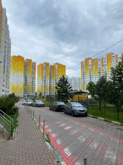 In the city of Goluboye, Russia, a residential street is depicted under an overcast sky. The street is paved with red and gray bricks, marked with faded red and white pedestrian crossing lines. Several cars are parked along the street, including a dark sedan, a silver sedan, and a blue hatchback. Tall, modern apartment buildings with bright yellow and white facades dominate the background. Green trees and a playground are visible to the right, separated from the street by a black metal fence. A metal handrail and walkway are visible on the left side of the frame.