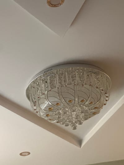 An ornate chandelier hangs from a white ceiling. The chandelier is silver and features numerous crystal droplets and amber-colored accents. It is suspended from a recessed ceiling. In the lower left of the image, a small, recessed circular light fixture is visible on the ceiling. The lighting in the image is soft and warm, suggesting it is daytime and interior lighting. The overall scene is clean and modern. The image is dated October 26, 2023, and the location is Yavoriv, Ukraine.