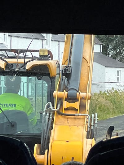 The image captures a close-up view of a yellow excavator. The operator, wearing a bright green high-visibility jacket with the text "TEASE CONSTRUCTION LTD" visible, is seated inside the cab. The excavator's arm is prominently featured, with its hydraulic hoses and mechanisms clearly visible. In the background, a row of white houses with dark roofs suggests a residential area. The presence of an excavator implies construction or maintenance work is underway. The overall setting appears to be outdoors, likely in a rural or semi-rural part of Portnablagh, Ireland, given the architectural style of the houses and the surrounding vegetation. The lighting suggests it is daytime.