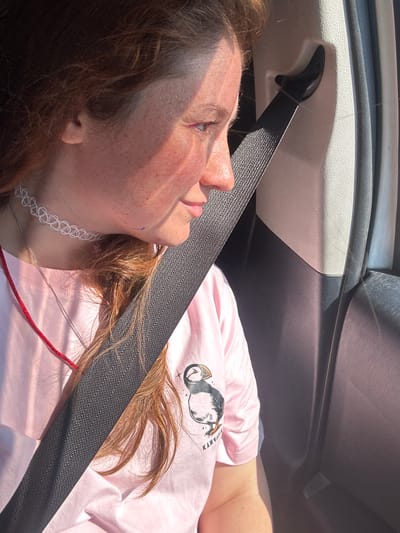A young woman with reddish-brown hair is shown from the chest up, seated in a vehicle. She is wearing a pink t-shirt with a puffin graphic and the word "KAMCHATKA" printed on it, a white choker necklace, and a silver chain. Sunlight streams in from the side, casting shadows across her face and the car's interior. A black seatbelt crosses her chest diagonally. The environment appears to be the interior of a car, possibly in or near the city of Krutoberegovoy, Russia, given the context provided.