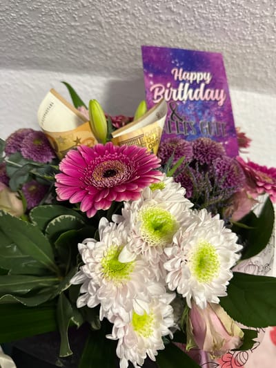 A bouquet of flowers is visible, containing pink gerbera daisies, white chrysanthemums with light green centers, purple pompom flowers, and Euro bills. The bouquet is accompanied by a "Happy Birthday & Alles Gute" card. The setting appears to be indoors against a plain, light-colored wall. Herxheim, Germany is not discernible from the image.
