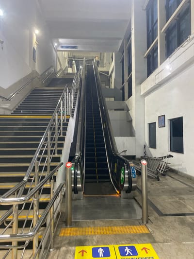 This media file captures a view of a public transportation station, likely a metro or train station, with escalators and staircases. The scene is well-lit with artificial lighting, suggesting it could be day or night. The setting appears to be indoors or partially covered, with concrete walls and ceilings, and metal railings.

The primary objects visible are two escalators, one going down and another going up, with their black steps and metal handrails. Alongside the escalators, there are metal staircases with yellow and black striped edges, indicating safety markings.

There are no people clearly visible in the frame, but the presence of escalators and signage suggests a public space with potential for transit.

Notable details include directional signage with text that is partially obscured but appears to include "Platform 1" and "Platform 2" with arrows. There are also yellow tactile paving tiles, commonly found in public spaces to assist visually impaired individuals. Additionally, a yellow sign with a red arrow and icons indicating accessibility for people with disabilities and general direction is visible.

The overall impression is one of a functional and modern transit hub. The orientation of the image is vertical, rotated 90 degrees counter-clockwise.