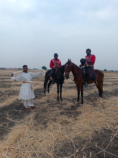 The scene is set in an open, harvested field in Kottalapalli, India, under an overcast sky. The ground consists of dark soil covered sparsely with light brown, dry crop stubble. Three adult males are present. On the left, a man stands facing the viewer, dressed in a white traditional Indian kurta and dhoti with a yellow waist sash, holding a white cloth extended horizontally with both hands. His feet are bare, with brown sandals visible beneath his attire. To his right, two men are mounted on horses. The man on the black horse, positioned slightly behind the other mounted individual, wears a red military-style jacket with white and black trim, black pants, black boots, and a black cap; he holds the reins. The man on the brown horse, positioned closer to the foreground on the right, also wears a similar red military-style jacket, black pants, and black boots. He holds a black hat in his left hand, and a sword or scabbard is visible attached to his left hip. Both horses are saddled and bridled, standing still. In the distant background, sparse trees and a flat rural landscape are visible.