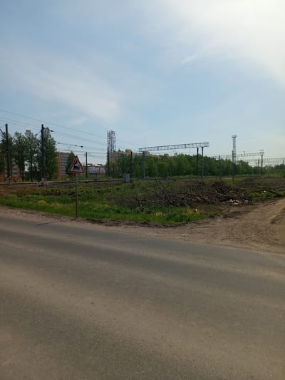 The image shows a street scene near a railroad track in Kommunar, Russia. In the foreground, there is an asphalt road. To the side, there is a grassy area with small yellow flowers. A triangular "railroad crossing" sign is visible. Behind the grass, a pile of tree branches lies near the railroad tracks. Metal structures and power lines are positioned above and along the tracks. In the background, apartment buildings are visible interspersed with trees, all under a pale blue sky with light cloud cover.