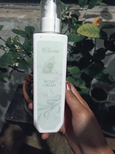 A white bottle of Olivia Levante body cream is held by a person's hand in front of a blurred background of greenery. The bottle has a light green label with text and an olive branch illustration. The background shows out-of-focus leaves and a potted plant. The scene is lit, with shadows cast on the hand and bottle. There is no discernible location in the city of Кош-Дөбө, Kyrgyzstan.