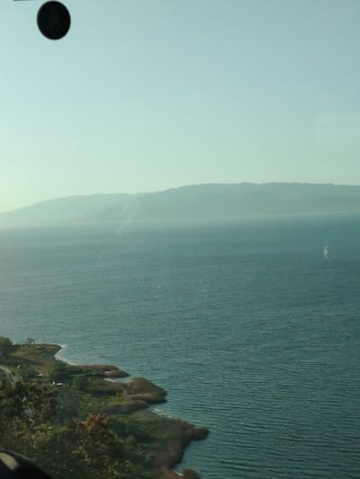The image captures a wide-angle view of a large body of water, identified as Lake Iznik in Turkey, under a clear, bright sky. A distant range of mountains, softened by haze, forms the horizon across the far side of the lake. The water itself appears a deep teal or greenish-blue, with small ripples visible across its surface, suggesting calm conditions.

In the foreground, a rugged coastline curves into the frame from the bottom left, featuring a mix of green foliage, trees, and patches of dry, reedy vegetation at the water's edge. Some very faint, small structures are discernable amidst the greenery on the near shore, and a hint of a road or path can be seen.

A notable feature is a dark, circular object positioned in the top left corner of the frame, appearing to be an artifact such as a reflection within glass or part of the camera's mechanism, with a faint, indistinct shape visible within it. Towards the middle-right of the lake, a faint, elongated white shape is visible, possibly a distant boat, a reflection, or an optical anomaly.

The lighting suggests it is daytime, possibly morning or late afternoon, with a soft, somewhat diffused light across the expansive scene. There are no visible people or discernible text in the image. The overall impression is one of natural serenity and vastness.