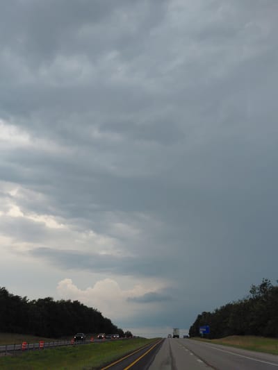 The media file captures a highway scene in Midway, United States of America, under a cloudy sky. The perspective is from the road, looking forward along the lanes. Trees line the right side of the road, with a grassy shoulder and a guardrail dotted with orange traffic cones.

Several vehicles are visible on the highway, including a truck in the distance and a few cars closer up. The headlights of some vehicles are illuminated, suggesting it may be late afternoon or early evening, or that the weather has reduced visibility. The sky is filled with large, dramatic clouds, hinting at potential impending rain or a storm. The overall mood is somewhat somber due to the overcast conditions.

A blue road sign with white text is visible on the right side of the highway, partially obscured by trees. While the text is blurry, it appears to be directional information or an exit marker. The road itself is paved and marked with yellow and white lines, indicating multiple lanes of travel. The presence of traffic cones suggests a possible construction zone or a temporary traffic diversion. The scene is characterized by the vast expanse of the sky meeting the linear path of the highway, bordered by natural vegetation.