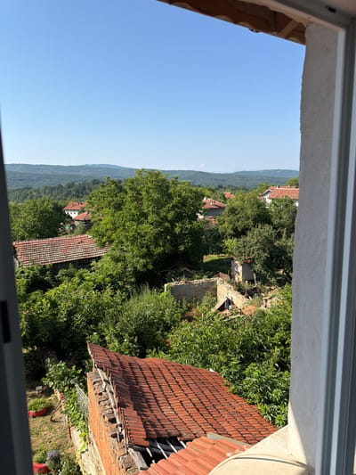 The image depicts a view through a window opening onto a rural landscape in the city of Gorno Pavlikene, Bulgaria. The scene is dominated by lush green trees and vegetation, with several buildings featuring red tile roofs visible amongst them. In the foreground, a partially constructed or dilapidated brick structure with a red tile roof is prominent. In the mid-ground, more houses with red tile roofs are scattered, some appearing well-maintained, while others, like the structure in the center, show signs of disrepair with exposed beams and rubble. The background reveals rolling hills covered in dense forests under a clear blue sky. The overall impression is that of a sunny day in a village setting. There are no people visible in the image.