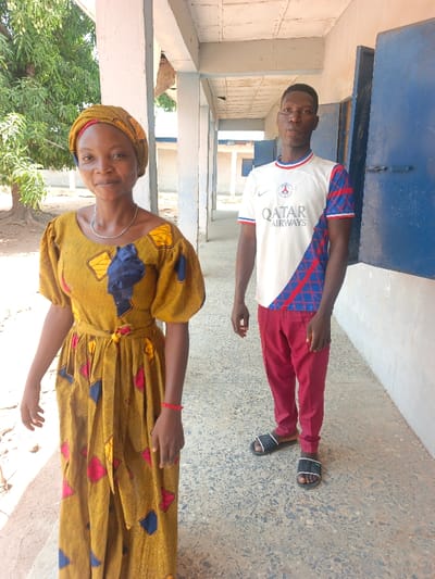 The image displays two individuals, one female and one male, standing on a paved outdoor walkway under a concrete overhang in the city of Mada, Nigeria. The female, positioned in the foreground to the left, wears a long, mustard-yellow patterned dress with puff sleeves, a matching headwrap, a thin silver-colored necklace, and a red wristband on her left arm. She is looking directly at the camera with a slight smile. The male, standing behind her to the right, wears a white short-sleeved t-shirt with "QATAR AIRWAYS" and a PSG logo, red trousers, and dark slide sandals. He is also looking directly at the camera with a neutral expression. The walkway features light-colored concrete pillars and a ceiling. To the right, a wall with open blue-painted shutters or doors is visible. The ground is a light-colored, speckled paved surface. In the background, green trees are present to the left, and other light-colored buildings with blue accents are visible under clear daylight. No overt interaction occurs between the individuals.