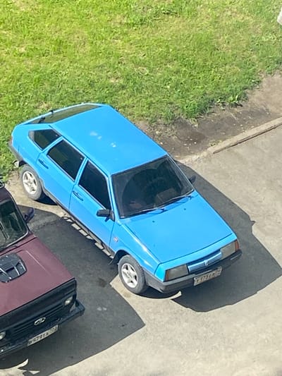 The image shows two parked cars on a paved surface next to a grassy area. The primary subject is a bright blue, five-door hatchback, viewed from a high angle. The car has tinted windows, silver alloy wheels, and a license plate visible on the rear. To the left and slightly in front of the blue car is the front section of a darker colored vehicle, possibly a maroon SUV or sedan. This second vehicle also has tinted windows and a license plate. The environment appears to be an outdoor urban setting, potentially a parking lot or residential area, with a patch of green grass to the upper left and a concrete or asphalt ground surface. The sun is casting shadows from the cars onto the ground. The overall impression is a still, everyday scene.