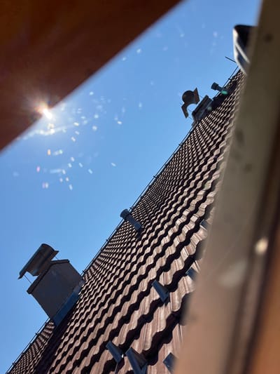 An angled, eye-level shot captures a tiled roof against a clear blue sky. The sun shines brightly, creating a lens flare effect with rainbow hues. Two chimneys are visible on the roof. One chimney on the left is square and dark-colored, topped with a silver, angled cap. A second, smaller chimney is further up the roof on the right, also dark-colored with a cylindrical vent. The roof tiles are reddish-brown and appear to be made of clay, arranged in a wavy pattern. The image was taken in Cacak, Serbia.