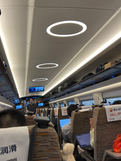 The image depicts the interior of a high-speed train carriage, likely in Tengzhou City, China. The ceiling features modern, elongated oval LED lights casting a soft glow, accentuating the curved lines of the carriage's structure. Along the sides, overhead compartments are filled with luggage, including backpacks and boxes. Passengers are seated in rows of brown and beige patterned seats. Several individuals are visible, including a man standing in the aisle wearing a grey t-shirt and glasses. Others are seated, with some engaged with laptops. Screens displaying what appear to be informational content or entertainment are mounted on the ceiling above the seats. The overall environment is clean and well-lit, suggesting a typical travel setting on a modern train.