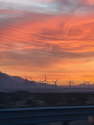 The image presents a striking sunset scene over a desert landscape, filled with wind turbines and observed from a vehicle. The sky dominates the upper portion, ablaze with vibrant hues of orange, pink, and yellow, transitioning to a softer grey-blue at the very top. Wispy, scattered clouds catch the warm, intense light of the setting sun.
Several dark power lines stretch horizontally across the sky in the upper-mid section, creating a visual frame. Below the colorful horizon, a vast array of wind turbines stands silhouetted against the twilight. Their sharp, dark forms are spread densely across the midground, suggesting an extensive wind farm nestled among rolling hills or mountains that recede into the distance with a hazy, dark profile. The foreground reveals what appears to be the top edge of a car's dashboard or window frame, indicating the photograph was taken from a moving vehicle.
The scene captures the notable details of a sunset, with its characteristic warm light and long shadows. The numerous wind turbines are a strong location cue, indicative of a region like Desert Hot Springs, United States, known for its wind energy installations. No people are visible, and there is no discernible text in the image. The overall impression is one of natural beauty intertwined with human engineering, under the majestic spectacle of dusk.