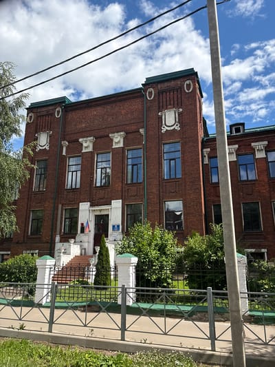 The image shows a brick building with white decorative elements and green trim under a bright blue sky with scattered clouds. The building appears to be an older structure, possibly a school or administrative building, given the signage visible. A metal fence runs along the foreground, separating a sidewalk from a grassy area and more of the building. Trees and greenery surround the building and fence.
On the building, there are several windows, some with reflections of the sky. One section of the building features a prominent entrance with white pillars and steps leading up to it. There are two flags visible near this entrance, one appearing to be the Russian flag. A blue sign with white text is mounted above the entrance.
The overall scene suggests a daytime setting on a pleasant day. The perspective is slightly tilted, making the image appear as if it's viewed from a low angle or that the scene itself is not perfectly level. The context indicates the location is Pereslavl-Zalessky, Russia. No specific activity or event is discernible; it appears to be a static view of the building and its surroundings.