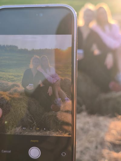 The image captures a "photo within a photo" scenario, where a smartphone is held up to photograph a scene, with its screen displaying the live camera feed. The primary focus is on the smartphone, likely an iPhone given its distinct notch/Dynamic Island design, held vertically by a hand partially visible in the bottom left corner.

On the phone's screen, two young women are depicted sitting closely on hay bales in an open field. The woman on the left, with blonde hair, wears a dark zip-up top and holds a white smartphone, looking directly at the camera with a serene expression. Her companion on the right, wearing a light-colored (possibly pink) jacket, has her arm around the first woman's shoulder and also looks at the camera with a slight smile. The background on the screen reveals a vast field with scattered hay bales, lush green grass, and a distant line of trees under a bright, warm sun, creating a significant lens flare and a soft, golden glow across the image.

Behind the phone, the actual scene being photographed is visible but intentionally blurred, emphasizing the phone's screen. The shapes of the two women, the hay bales, and the intense golden light are discernible, mirroring the content displayed on the screen.

The time of day is clearly the golden hour, either sunrise or sunset, indicated by the intense warm light and the sun's low position, which creates the prominent lens flare. The setting is an outdoor, rural landscape, specifically located in Стеклянный, Russia, characterized by agricultural fields and natural elements. The activity is the act of taking a photograph, capturing a moment of friendship and relaxation in nature.

Visible text on the phone's camera interface includes zoom options "0.5", "1", "2x", "5" (with "2x" highlighted), the word "PHOTO" indicating the mode, a circular shutter button, "Cancel" option, and a camera-switch icon.