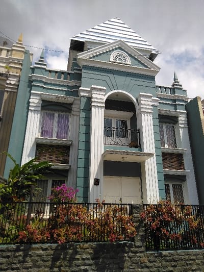 The image depicts a multi-story residential building located in Nathene, Laos, viewed from a slightly low angle on a partially cloudy day. The structure features a predominantly light teal-green facade contrasted with white architectural trim, columns, cornices, and window frames. A prominent, multi-tiered pyramidal roof, appearing silver or light grey, crowns the building.
The central portion of the upper facade includes a large arched recess containing a balcony with a black wrought-iron railing, accessible via double glass doors. This arch is flanked by decorative white fluted columns. Above this, a white pediment features a semicircular window with radial spokes. On either side of the central arch, the upper story presents windows with white frames; the left window has purple translucent curtains.
The ground level features a large, double white entrance door, recessed beneath a small white overhang. To the immediate left of this entrance, a black rectangular plaque displays the numbers "315 / 1B". Windows are present on both sides of the ground-level door.
The property is fronted by a low stone wall, topped with a black wrought-iron fence. Over and through this fence grows a dense arrangement of plants, primarily featuring reddish-brown foliage and a cluster of vibrant purple flowers on the left side. Additional green foliage is visible within the property behind the fence. Sections of adjacent buildings are discernible on both the left and right edges of the frame.