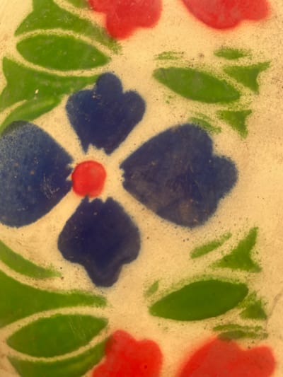 This image presents a close-up, top-down view of a decorative surface located in Awgbu, Nigeria. The surface, appearing light off-white or cream-colored, is adorned with a floral-themed pattern executed in dark blue, red, and green paint or enamel.
The central element is a four-petal flower design, featuring a small, circular red center from which four dark blue, rounded, petal-like shapes extend radially. Surrounding this central motif are numerous green, elongated, leaf-like forms, often arranged in pairs or clusters. Additionally, several rounded red shapes are visible at the edges of the frame, suggesting parts of other floral elements or a continuous pattern extending beyond the captured area.
The painted application exhibits a textured appearance, with visible slight wear, minor abrasions, and scattered dark specks across the surface, particularly within the blue and green areas, indicating age or exposure. No individuals, specific objects beyond the painted surface, actions, or interactions are present or discernible within the frame. The environment is limited to the close-up of this patterned surface.