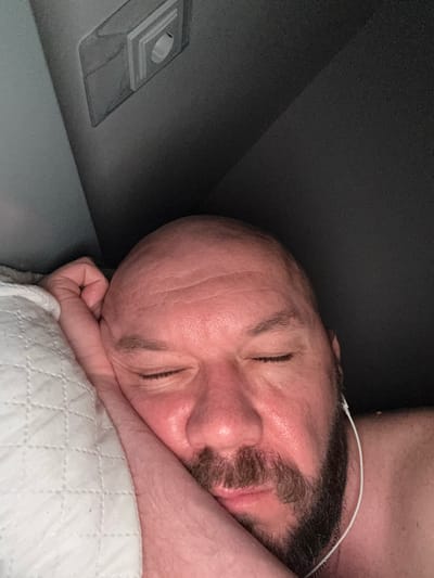 A close-up, overhead shot shows a bald, middle-aged man with a beard sleeping on his side, resting his head on a white quilted pillow. His eyes are closed, and his face is flushed. He is wearing white earbuds. The background consists of a dark, textured surface, possibly the interior of a vehicle, with a light fixture visible in the upper left corner. The scene is captured indoors in Starychi, Ukraine.