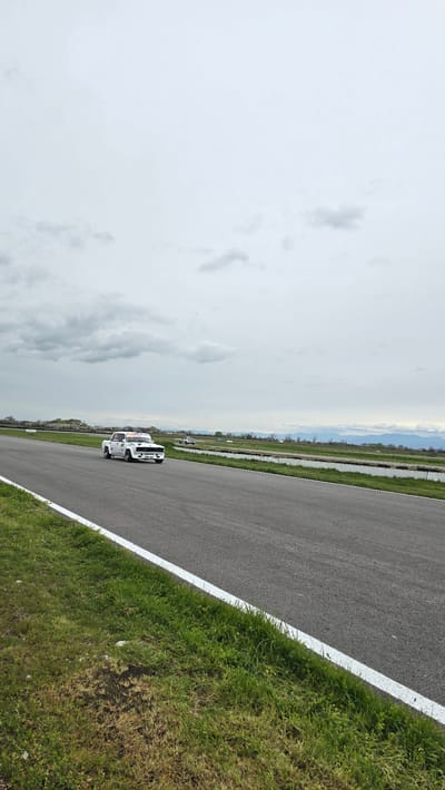 The image shows a racetrack in Kaloyanovo, Bulgaria. Two white rally cars are driving on the asphalt track. The track has a white line on its right edge, bordering green grass. The sky is cloudy. In the distance, there are trees, land, and mountains.