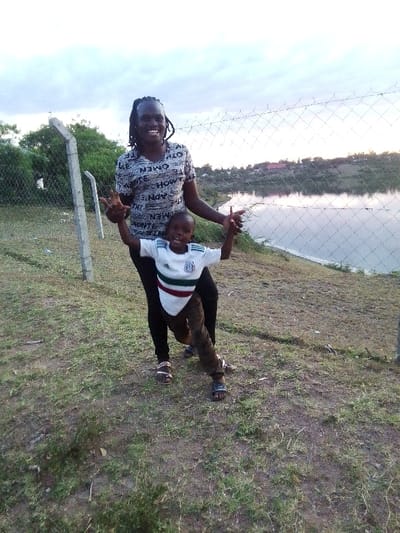 An adult woman and a young boy are captured in a joyful outdoor scene in Central Kasipul ward, Kenya. The woman, with dark skin and dreadlocks, wears a light grey t-shirt featuring repeated text patterns like "MEN," "WOMEN," "HOW," and "THE," along with dark pants and sandals. She is smiling broadly and holding the hands of the young boy, who also has dark skin and a wide smile. The boy wears a white t-shirt with distinct horizontal red, white, and green stripes, patterned brown pants, and sandals.
They are standing on a patch of dry grass and dirt, positioned in front of a chain-link fence with concrete posts. Beyond the fence lies a calm body of water, likely a lake or reservoir, reflecting the soft light of the sky. The far bank is covered with green vegetation and shows subtle hints of distant buildings or settlements. The sky is bright but suggests either an overcast day or the soft light of late afternoon or early evening. Both subjects exhibit clear happiness, indicating a warm and playful moment between them.